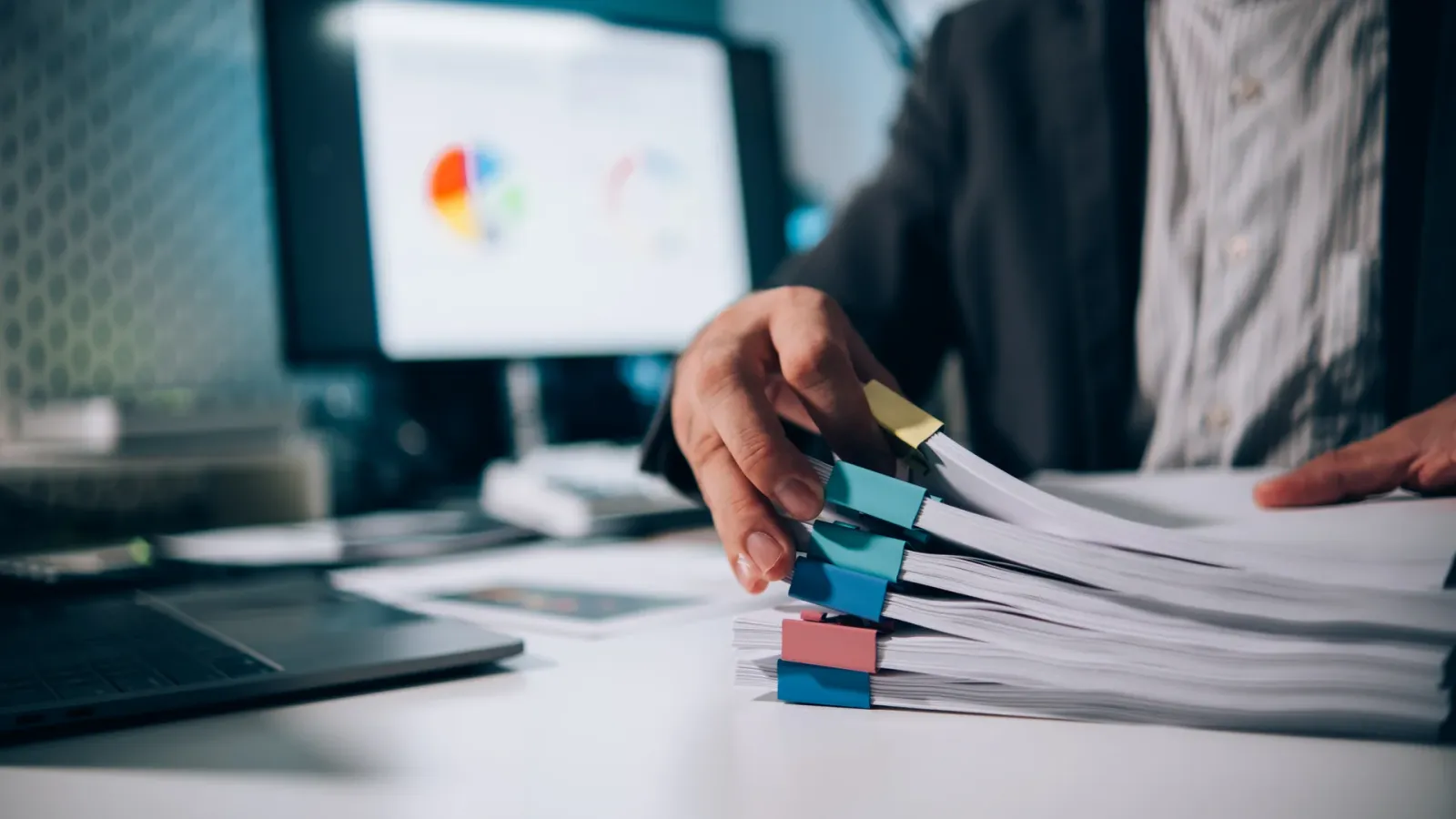 Hands sorting a stack of paperwork in front of a screen showing charts