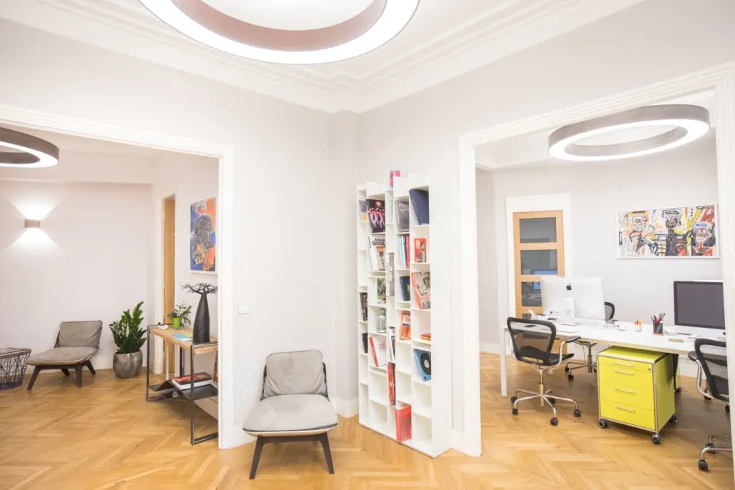 Les bureaux de Paradigm : parquet à chevrons, moulures, luminaires circulaires, postes de travail et coin salon séparés par une bibliothèque ouverte.
